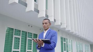 A professional man in a blue suit reviews documents on a laptop outside a modern building. video