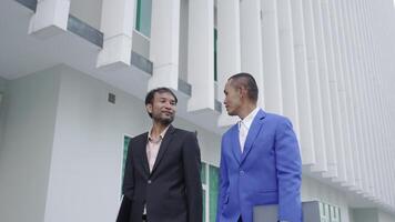 dos empresarios en formal trajes caminar juntos fuera de un moderno edificio, que lleva carpetas el pulcro arquitectura en el antecedentes sugiere un profesional o corporativo ajuste. video