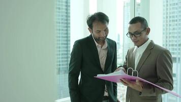 dos empresarios en formal atuendo estar por un oficina ventana con vista a el ciudad, revisando documentos. uno hombre gestos con un Pulgares hacia arriba, indicando aprobación o acuerdo. video