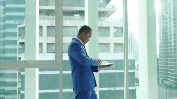 A businessman in a blue suit works on his tablet near a window overlooking the city. Another professional walks by, highlighting a dynamic office environment. video