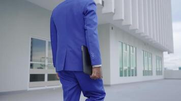 A close-up of a businessman in a blue suit holding a laptop as he walks outside a modern office building. The scene emphasizes professionalism, technology, and corporate ambition. video