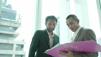 A businessman holding folders approaches a colleague who is standing by a large window, gazing out at the city skyline. The scene conveys reflection and corporate planning. video