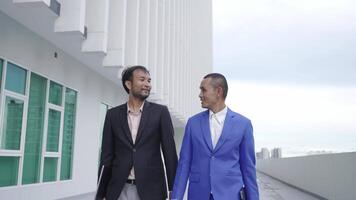 Two businessmen in formal suits walk together outside a modern building, carrying folders. The sleek architecture in the background suggests a professional or corporate setting. video