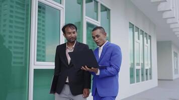 Two businessmen stand outside a modern office building, reviewing information on a laptop. One man explains something while the other listens attentively, reflecting teamwork and collaboration. video