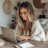 Smiling young woman with glasses working on a laptop in a cozy home office, surrounded by minimalist decor and notebooks photo