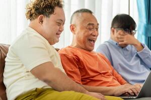 Senior asian man having a good time sitting on couch at home with adult son, Cheerful grandfather and grandson using laptop doing a call in living room photo