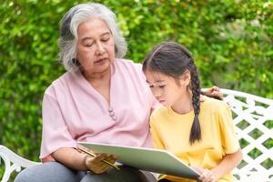 Senior Asian woman and her granddaughter play together in the garden, Grandmother with kid girl drawing picture in the yard photo
