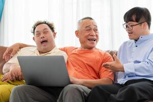 Cheerful grandfather and grandson using laptop doing a call in living room, Senior asian man having a good time sitting on couch at home with adult son photo