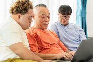 Grandfather and grandson using laptop doing a call in living room, Asian senior man having a good time sitting on couch at home with adult son photo