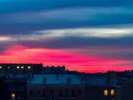 A stunning sunset scene with deep pink and blue hues in the sky, silhouetting city rooftops. A few lit windows in the darkening buildings add warmth and contrast to the vibrant evening sky photo
