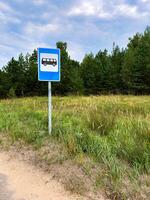 A bus stop sign stands on the side of a quiet rural road, surrounded by grassy fields and a backdrop of dense pine trees. The blue sky with scattered clouds adds a peaceful atmosphere to the scene photo