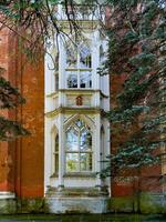 A close up of a beautifully preserved Gothic window from a historic estate, framed by red brick and lush pine branches. The intricate architectural details and glass reflections emphasize the building photo