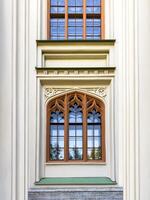A close up of a beautifully preserved Gothic window from a historic estate, framed by red brick and lush pine branches. The intricate architectural details and glass reflections emphasize the building photo