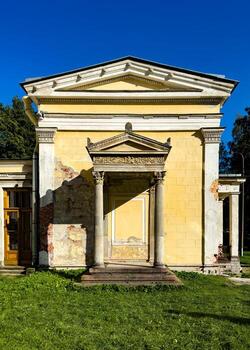 Front view of a weathered portico with classical columns and intricate stone carvings on the facade of an abandoned villa. The faded yellow walls show signs of decay, adding a touch of historical photo
