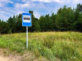 A bus stop sign stands on the side of a quiet rural road, surrounded by grassy fields and a backdrop of dense pine trees. The blue sky with scattered clouds adds a peaceful atmosphere to the scene photo