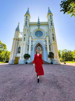 A woman dressed in a flowing red gown walks confidently in front of a grand Gothic-style cathedral. The ornate architecture of the building, with its tall spires and intricate details, creates a photo