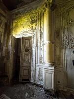 A detailed shot of the crumbling interior of an abandoned palace, revealing ornate plasterwork, decorative columns, and a weathered wooden door. Sunlight filters through, casting an eerie glow on the photo
