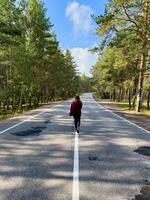 A carefree woman walks down an empty country road lined with pine trees, arms outstretched in the wind. Captured from behind, the scene emphasizes the wide open space, cracked asphalt, and serene photo