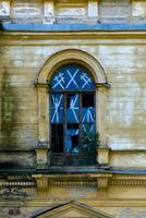 A haunting image of a broken arched window in an abandoned mansion. The peeling yellow paint of the building contrasts with the shattered glass panes and crisscrossing tape. Overgrown plants peek photo