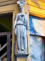 A close-up view of a classical female statue mounted on the corner of a historic building, illuminated by warm sunlight. The weathered stone figure contrasts with the vibrant yellow facade photo