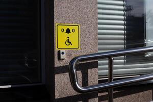 Grasping railings on handicapped ramp next to disabled person in wheelchair sign on wall of public building with button to call help, equipped entrance with ramp for disabled people photo
