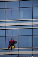 Industrial climber at difficult job, vertical photo of climber washing windows of glass skyscraper, cleaning exterior of modern building