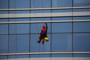 Worker in uniform cleans glass of skyscraper while hanging on ropes, difficult job of climber with risk to his life when he cleaning windows of building photo