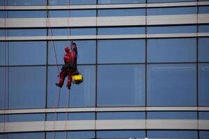 Industrial climber in red overalls cleans the glass windows of skyscraper with office space, hard work at height for cleanliness of building exterior photo