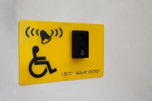 Yellow sign on wall of building with sign of person in wheelchair next to button to call help and braille text for blind people. Helping people with disabilities on the street in city, accessible environment photo