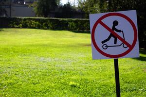 Sign in grassy field says no scooter in park. Sign is red and black. Symbol prohibiting the use of scooter near field green in background photo