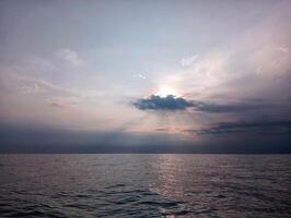 A large body of water with a sun shining through the clouds photo