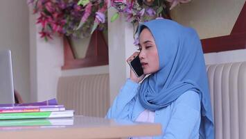 Beautiful asian woman hijab is focusing on calling her friend using mobile phone in front of laptop on table in coffee shop. for communication, advertisement, lifestyle, entertainment, business video