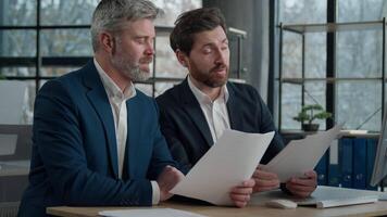Two Caucasian men businessmen in office working together on financial statistics analyzing charts project discussing data in report doing paperwork using computer looking documents client consultation video