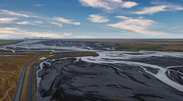 A stunning aerial view of a wide, shallow river winding through black sand in Iceland. A road runs along the left side, with a blue sky and white clouds above. photo