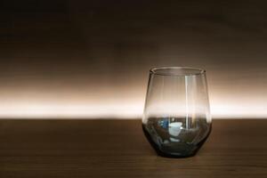 A single, empty glass cup sits on a dark wooden table. The glass has a curved bottom and a wide rim. The background is a gradient of dark brown to a lighter brown. photo