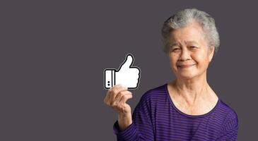 Service rating, satisfaction concept. Portrait of an elderly Asian woman holding a thumbs up a symbol and looking at the camera with a smile while standing on a gray background photo