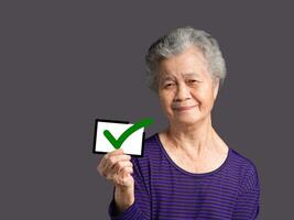 Senior woman holding a thumbs up a symbol and looking at the camera with a smile while standing on a gray background photo