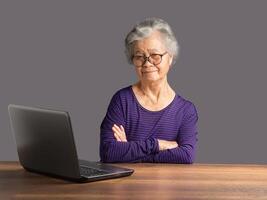 An elderly Asian woman with short gray hair sitting with arms crossed, looking at the camera and a laptop on the table. photo