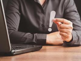 Exclamation mark concept. Hand of a businessman in a black shirt holding a white exclamation mark while sitting in the office. photo