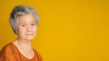 An elderly Asian woman with short gray hair looking at the camera with a smile while standing on a yellow background photo