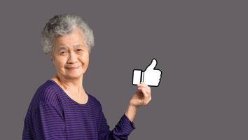 An elderly Asian woman holding a paper thumbs up and looking at the camera with a smile while standing on a gray background photo