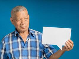 Senior man holding and looking at a blank paper speech bubble while standing on a blue background photo