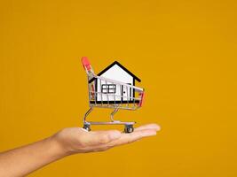 Home loan and real estate investment concept. Close-up of a paper white house in the mini shopping trolley on the palm while standing on a yellow background photo