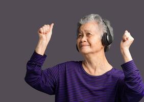 Cheerful elderly Asian woman wireless headphones to listen to a favorite song with a smile while standing on a gray background. photo