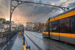 A tram traverses the iconic Dom Luis I Bridge in Porto, Portugal, during a dramatic sunset. The wet surface and historic buildings create a reflective, atmospheric scene. photo