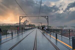 Dramatic sunset over Dom Luis I Bridge in Porto, Portugal, with tram tracks, pedestrians, and the Serra do Pilar Monastery in the background. Cloudy sky adds atmosphere. photo