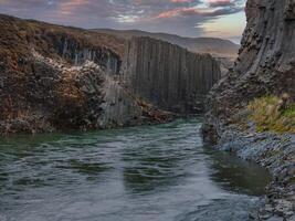 A calm green river meanders through a narrow basalt canyon with steep dark gray walls under an overcast sky in Iceland. The light is soft and diffused. photo