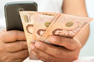 Asian woman holding Thai banknotes money with smartphone, economy finance exchange trade investment. photo