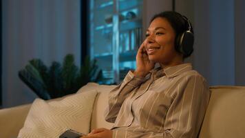 Smiling African American woman in headphones on couch at evening listen song home leisure with modern tech female music lover listening mobile phone audio girl relaxing on couch in night living room photo