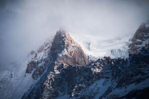 Majestic Mountain Peak emerging through Mist and Clouds, with a combination of Snowy Ridges and Rugged Rock formations. Interplay of Light and Shadow highlights the Dramatic natural beauty photo